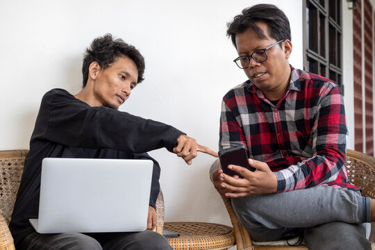 Two Indonesian southeast asian men using a laptop and a cellphone to collaborate while sitting at home. Discussing something on the screen of the mobile device. Work from home