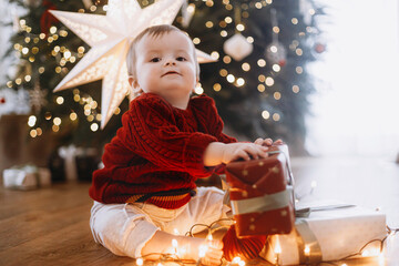 Merry Christmas and Happy Holidays! Cute baby in knitted sweater sitting with stylish gift box and lights at decorated christmas tree and star. Atmospheric magical moment