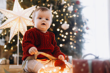 Merry Christmas and Happy Holidays! Cute baby in knitted sweater sitting with stylish gift box and lights at decorated christmas tree and star. Atmospheric magical moment