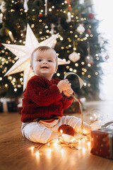 Merry Christmas and Happy Holidays! Cute baby in knitted sweater sitting with stylish gift box and lights at decorated christmas tree and star. Atmospheric magical moment