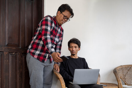 Two Indonesian southeast asian men using a laptop and a cellphone to collaborate. One sitting with a laptop and the other standing with a mobile device. Work from home