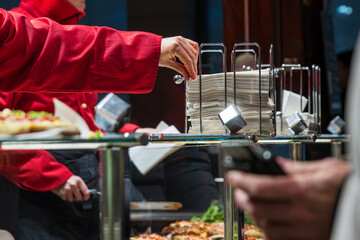 Vendor in a red jacket pulling napkins from a metal dispenser, serving food trays for customers in...