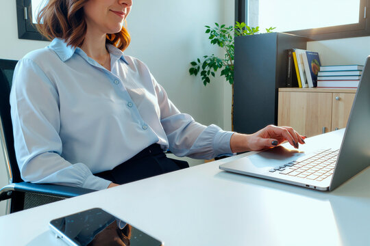 Caucasian young adult woman sitting at desk working on laptop, hand on laptop touchpad, smartphone on table, bookshelf and plant in background, business office environment