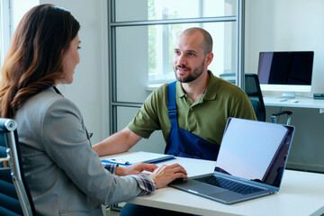 Caucasian young adult man with beard sitting across from Caucasian young adult woman discussing work at desk with open laptop in modern office setting