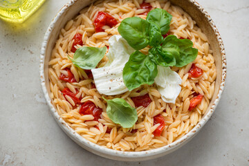 Orzo with torn mozzarella, roasted cherry tomatoes and fresh green basil, horizontal shot on a light-beige stone background, middle close-up
