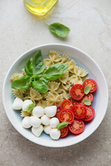 Plate of caprese pasta salad with mini mozzarella, cherry tomatoes, fresh basil and farfalle, vertical shot on a beige stone background