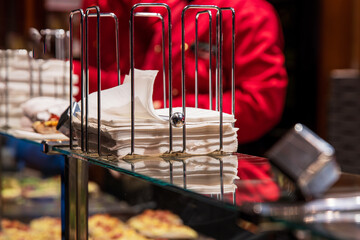 Winter street food stall, showing a neatly arranged stack of white napkins in a metal holder, with a vendor in red uniform working behind the counter. 
