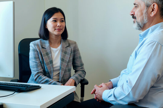 Asian young adult woman listening attentively to Caucasian middle aged man during business meeting in modern office setting, both seated near computer monitor and keyboard