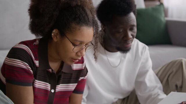 Biracial couple sitting together at home reviewing household bills and calculating expenses, managing finances in realistic and relatable domestic setting
