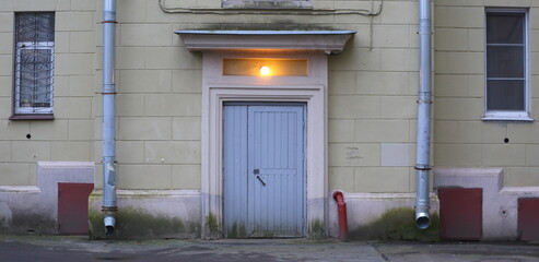 The wall of an old house with narrow windows, drainpipes, and a burning lamp above the wooden front door, Tallinn Street, Saint Petersburg, Russia, November 27, 2025