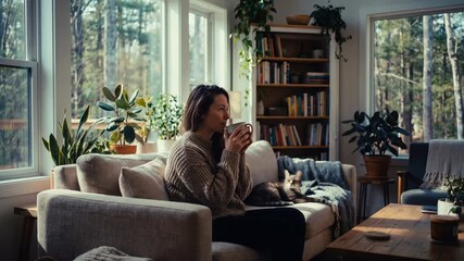 Young woman relaxing with hot drink on cozy sofa in sunlit living room, morning self care and mindful routine with houseplants and cat companionship for wellness and home comfort concepts
