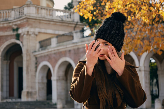 Mujer joven con abrigo marr&oacute;n y gorro de invierno  en un entorno de parque con &aacute;rboles de oto&ntilde;o