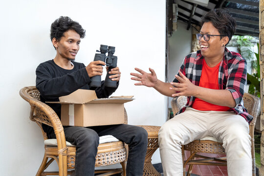 Two Indonesian southeast asian men sitting on a chair. Holding a package of a new pair of binoculars and showing it to his friend. Delivery service - Powered by Adobe