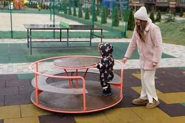 A child in a winter outfit spins on the merry-go-round at a park. A parent watches nearby, ensuring their safety while enjoying the cool day outdoors