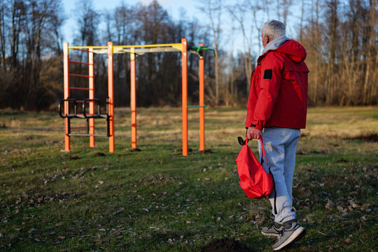 A man in a red jacket carries a bag as he approaches workout bars in a grassy park surrounded by bare trees on a clear morning