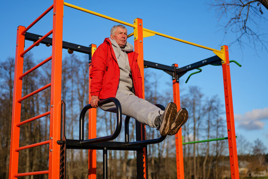 A senior man performs a dip exercise on bright orange fitness equipment in a park. Trees surround the area under a clear blue sky, showcasing an active lifestyle