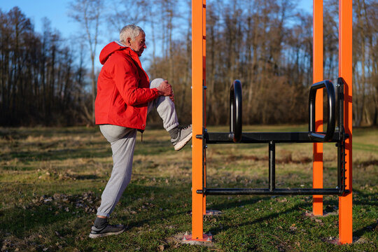 An older man in a red jacket stretches his leg at an outdoor fitness station in a park, surrounded by trees and sunlight during a clear day