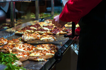 Hot cheesy flatbreads steaming in a tray while a vendor in gloves prepares more dough rounds topped with chopped tomato and slices of cheese. Festive street food preparation