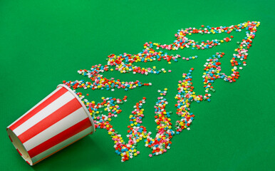 Multicolored sugar stars in the shape of a Christmas tree are scattered across a green background. A paper cup is also placed on the green background