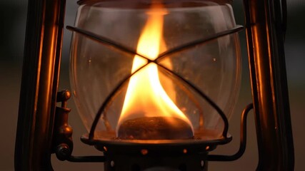 A close-up view of a burning lamp, with a vibrant flame inside the glass. The lamp structure is dark, creating a beautiful light