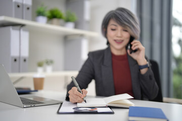 Older Asian businesswoman managing business writing notes making phone call modern office desk blurred professional multitasking busy work environment