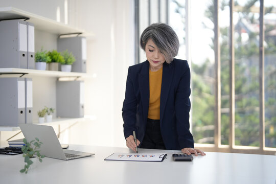Older Asian businesswoman analyzing financial reports charts data using calculator pen modern bright office workspace professional business management