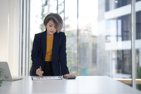 Asian older businesswoman analyzing financial data planning strategies using documents calculator pen modern office environment professional expertise business growth