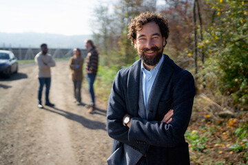 Smiling man stands confidently on a rural path while friends chat in the background during a sunny day in autumn