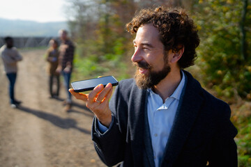Man using smartphone with voice command on a sunny day in a forest with friends in the background