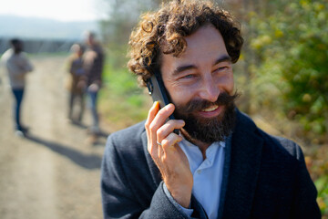 Man with curly hair talks on phone while people walk in background on a sunny day outdoors