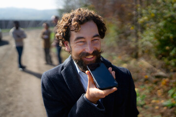 Happy man using smartphone to talk in green outdoor setting with friends in background during sunny day