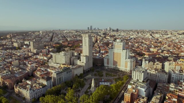 Cinematic ascending aerial view of Madrid, Spain, showcasing the iconic Plaza de Espa&ntilde;a, Edificio Espa&ntilde;a, and the distant Four Towers during a warm sunset.