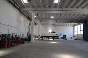 The interior of a large, with high ceilings and white metal beams. In the center, a medium-sized motorboat rests on metal trestles that lift it off the ground for maintenance or storage.