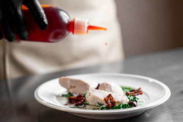 A chef in black gloves meticulously plates a gourmet chicken dish with creamy Tuscan sauce, spinach, and sun-dried tomatoes in a professional kitchen setting