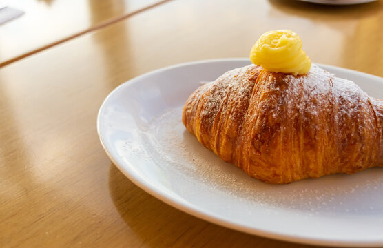 Primer plano macro de croissant o medialuna horneada, cubierta con copo de crema pastelera y az&uacute;car impalpable, servida en plato blanco sobre mesa de madera.