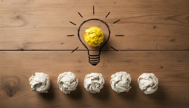 A bright yellow crumpled paper ball inside a drawn lightbulb, surrounded by five white crumpled paper balls on a wooden background, symbolizing new ideas and creativity.