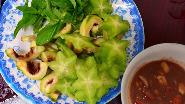 A plate of sliced green figs and sour starfruit with fresh herbs, served alongside a bowl of spicy fermented dipping sauce.