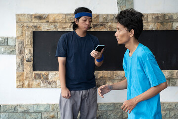 Two men are standing in front of a wall, one of them is holding a cell phone