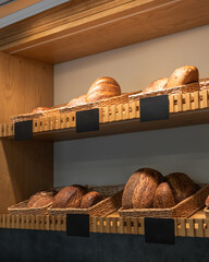 Artisanal bread display in bakery. Bread loaves displayed in wicker baskets on wooden shelves in a bakery. Several blank black labels are visible for pricing.