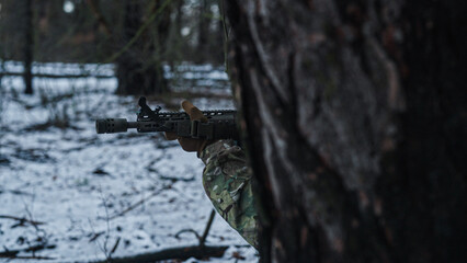 Soldier on a recon mission in the wilderness. Forest at winter. Aiming from behind a tree. War tactics