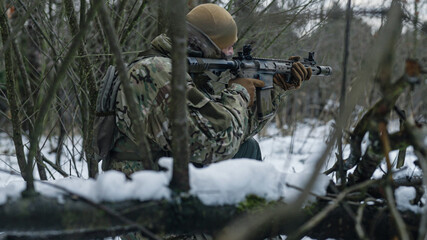 Soldier on a military recon mission in the wilderness. Forest full of snow at winter. Aiming  crouching on the ground. War tactics