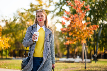 Confident businesswoman in stylish autumn coat holding laptop bag and drinking coffee while walking through city park during sunny day.