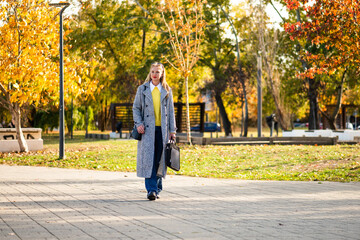 Urban businesswoman wearing sunglasses and stylish autumn coat while walking with a laptop bag through the city park during sunny day.