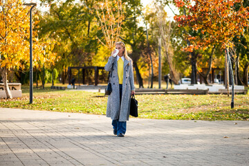 Stylish businesswoman in autumn coat holding laptop bag and drinking coffee while walking through city park during sunny day.
