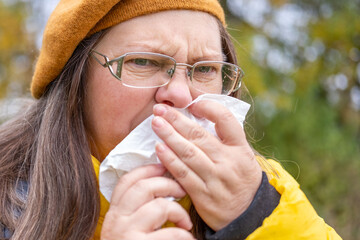 Sick mature woman with tissue, blowing nose, suffering from seasonal allergy, outdoor portrait, cold symptoms, sneezing, fatigue