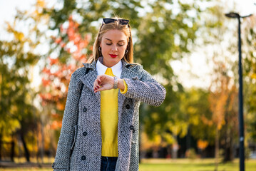Stylish businesswoman with sunglasses in modern autumn coat using smartphone while walking through the city park during sunny day.