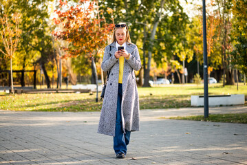 Confident businesswoman with sunglasses in modern autumn coat using smartphone while walking through the city park during sunny day.