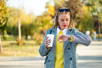 Confident businesswoman with sunglasses in stylish autumn coat checking the time on her wristwatch and drinking coffee while walking through the city park during sunny day.