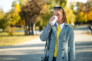 Portrait of urban businesswoman with sunglasses in stylish autumn coat drinking coffee while standing in the city park during sunny day.