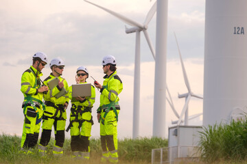 Team of wind energy technicians discussing project near wind turbines in green field during late afternoon